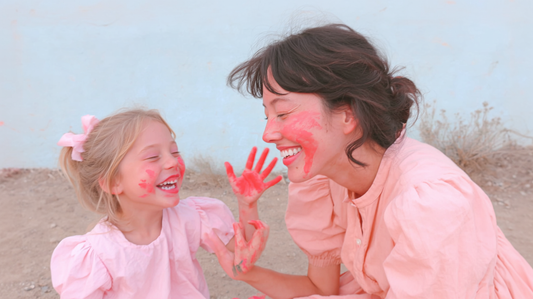 Mother and daughter laughing together with pink paint on their faces and hands, enjoying a playful painting moment outdoors.