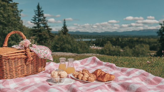 A pink picnic blanket with croissants, pastries, juice bottles, and a wicker picnic basket set on a grassy hill overlooking a scenic forest and mountains.